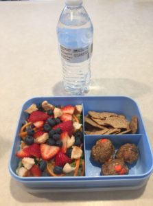 Blue lunchbox with berry salad which is made of greens, croutons, blueberries, and cut strawberries. Served with whole wheat crackers, energy balls, and water.