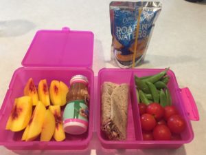 Pink lunchbox with peaches, whole wheat wrap, snap peas, and tomatoes. Drink is a Capri Sun.